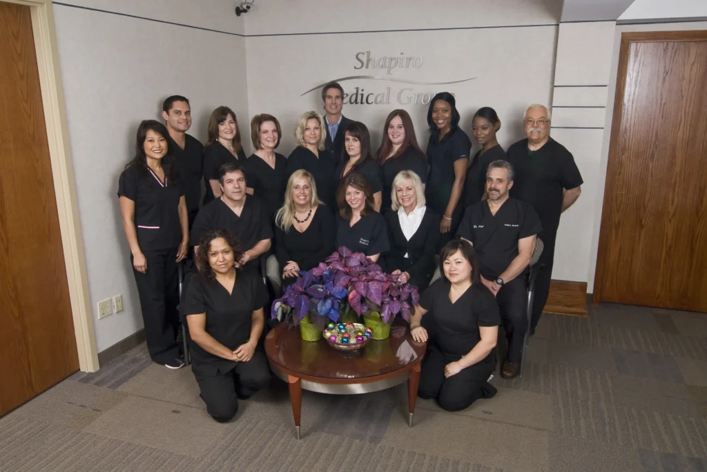 A diverse team of 17 healthcare professionals poses together in a well-lit office environment, wearing black uniforms. They are arranged in two rows around a table adorned with a colorful floral arrangement. The background features a logo or sign that suggests a medical or wellness practice. This image represents teamwork and professionalism in a healthcare setting, highlighting the staff's commitment to patient care.