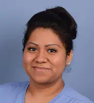 Portrait of a smiling healthcare professional wearing a blue scrub top, with dark hair styled in a bun. The background is a soft blue, creating a calm and professional atmosphere. This image represents the dedication and compassion found in the healthcare field, highlighting the importance of medical staff in providing quality care. Ideal for use in articles or websites focused on healthcare services, nursing, or medical education.