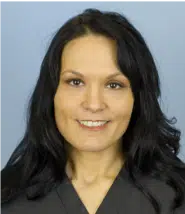 Professional headshot of a woman with long, dark hair and a friendly smile, wearing a gray blazer against a light blue background. This image represents a confident and approachable professional, suitable for business profiles, online portfolios, or corporate websites. The clear facial expression and neutral background enhance visibility, making it accessible for individuals with visual impairments.