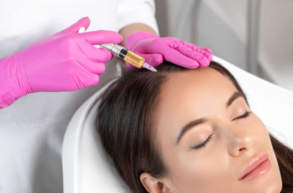 A healthcare professional wearing pink gloves administers a hair restoration treatment to a relaxed female patient. The procedure involves injecting a serum into the scalp to promote hair growth. The setting is a modern clinic, emphasizing a clean and professional environment. This image highlights the growing trend of non-surgical hair restoration therapies aimed at addressing hair thinning and loss.