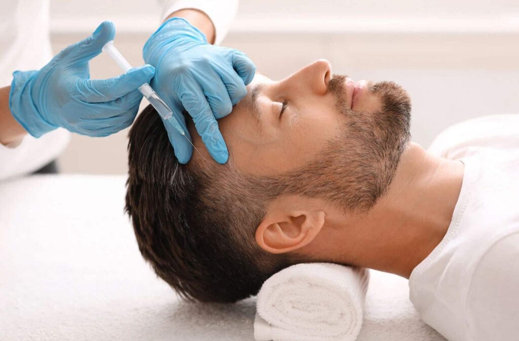 A male client receives a cosmetic treatment in a spa setting. A professional wearing blue gloves administers a facial injection, focusing on the forehead area. The client is relaxed, lying on a treatment table with rolled towels supporting his head. Soft natural light illuminates the room, creating a calming atmosphere. This image illustrates the growing trend of aesthetic procedures for men, highlighting skincare and wellness in modern beauty practices.