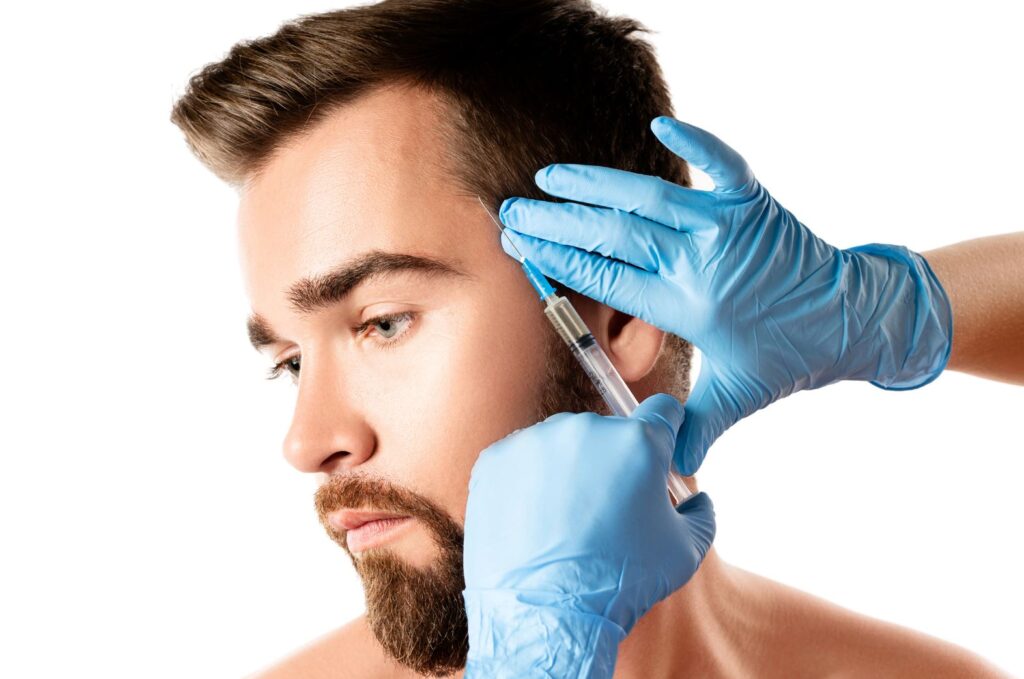 A professional administering a cosmetic injection to a young man’s jawline. The injector is wearing blue latex gloves, ensuring hygiene and safety. The focus is on the procedure, highlighting the growing trend of cosmetic enhancements among men. The young man appears calm and relaxed, emphasizing a positive experience. This image represents advancements in aesthetic treatments and the importance of professional care in cosmetic procedures.