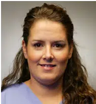 Portrait of a healthcare professional with long, wavy brown hair, smiling confidently. She is wearing a light blue medical scrubs top, suitable for a clinical or hospital setting. The neutral background emphasizes her friendly demeanor, making this image ideal for healthcare-related content. The expression and attire suggest professionalism and compassion, aligning with the values of patient care and support. This image can be used in articles or websites focusing on healthcare services, nursing, or medical professions.