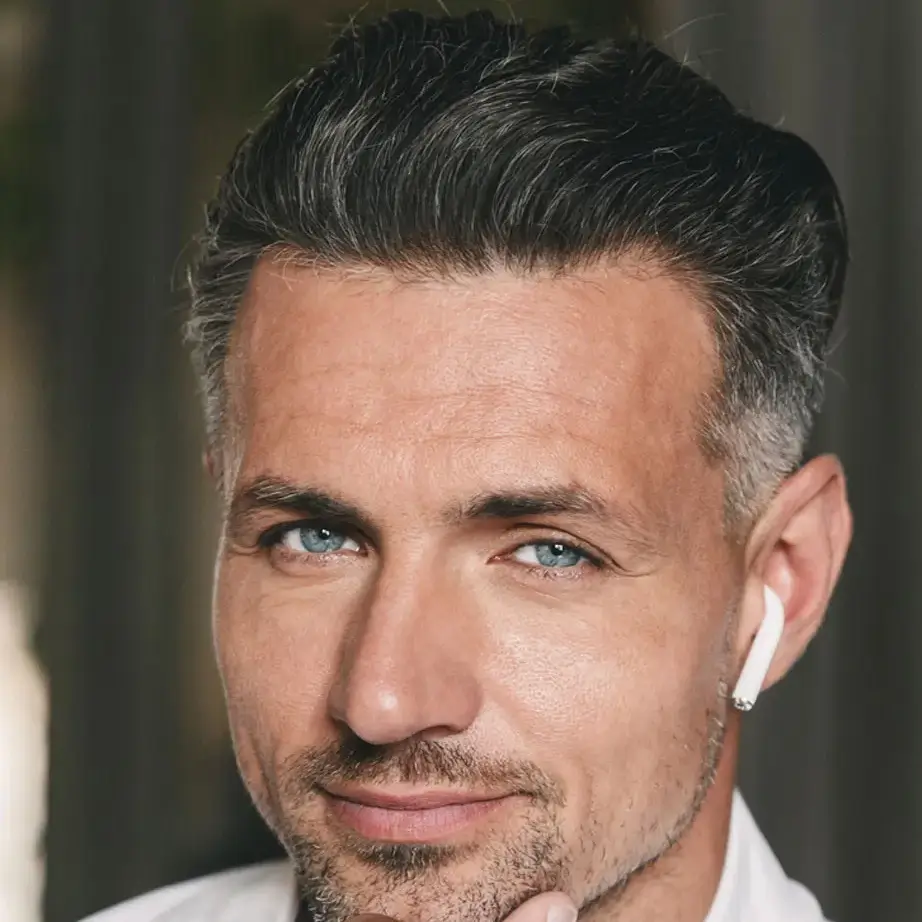Portrait of a man with short, stylish gray hair and a well-groomed beard, wearing wireless earbuds. He is looking directly at the camera with a thoughtful expression, set against a softly blurred background. This image conveys a modern and professional vibe, suitable for topics related to technology, lifestyle, or personal branding.