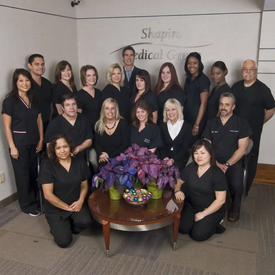 Group photo of a diverse team of 16 healthcare professionals wearing black scrubs, standing and sitting around a table with a vibrant floral arrangement. The background features a logo and the name of the clinic, emphasizing a welcoming and professional environment. This image highlights teamwork and expertise in providing quality healthcare services.