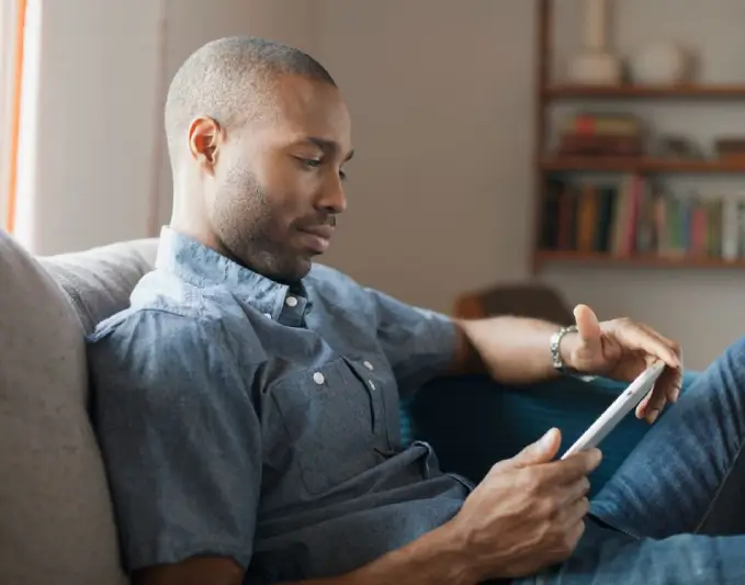 A man sitting comfortably on a couch, focused on reading a tablet device. He is dressed in a casual blue shirt and has a short haircut. The background features a cozy living room setting with bookshelves and soft lighting, creating a relaxed atmosphere. This image illustrates modern technology use in a home environment, highlighting the integration of digital devices in daily life. Ideal for discussions on technology, reading habits, or home decor.