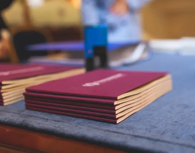 Stack of maroon presentation folders on a blue table, with a blurred background suggesting a professional setting. The folders are neatly arranged, showcasing their smooth edges and embossed logos. A blue stapler is visible in the background, indicating a workspace for organizing materials. This image conveys a sense of professionalism and preparation, suitable for business meetings or presentations. Ideal for articles about office organization, business branding, or presentation tips.