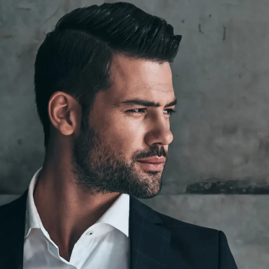 Stylish young man with short, well-groomed hair and a neatly trimmed beard, wearing a dark suit and white shirt. He is positioned against a textured gray background, exuding confidence and sophistication. The image captures a side profile, highlighting his strong jawline and thoughtful expression. Ideal for fashion, men's grooming, or professional attire content, this image conveys elegance and modern style.