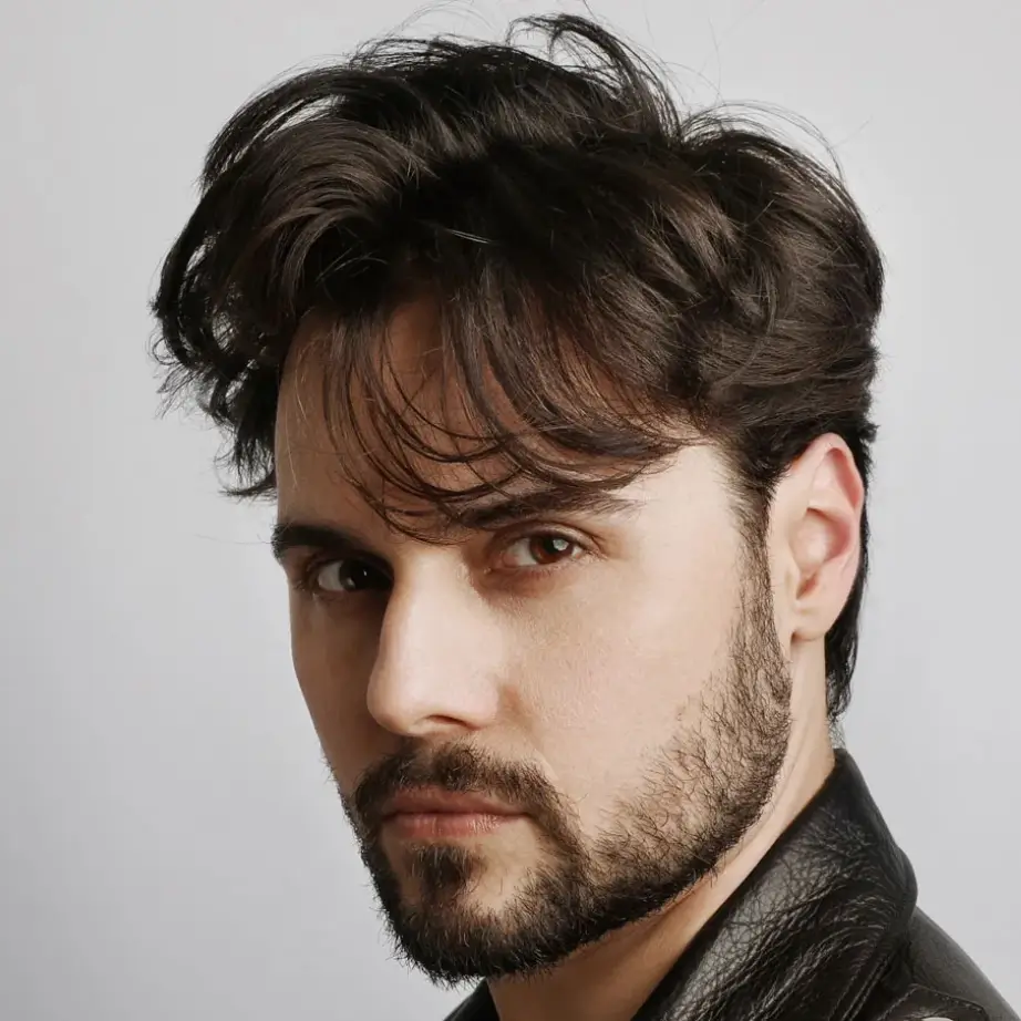 Portrait of a young man with tousled dark hair and a well-groomed beard, wearing a black leather jacket. He is facing slightly to the left, with a neutral expression against a light gray background. The image captures a stylish and modern look, suitable for fashion or lifestyle contexts.