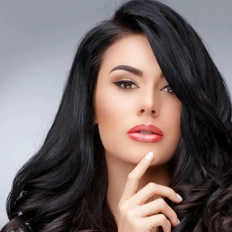 Portrait of a woman with long, flowing black hair and a subtle smile, gently resting her finger on her chin. She has striking features, including full lips and well-defined cheekbones. The background is softly lit, enhancing her facial expression and creating a professional and elegant atmosphere. This image is suitable for beauty, fashion, or lifestyle content, emphasizing confidence and poise.