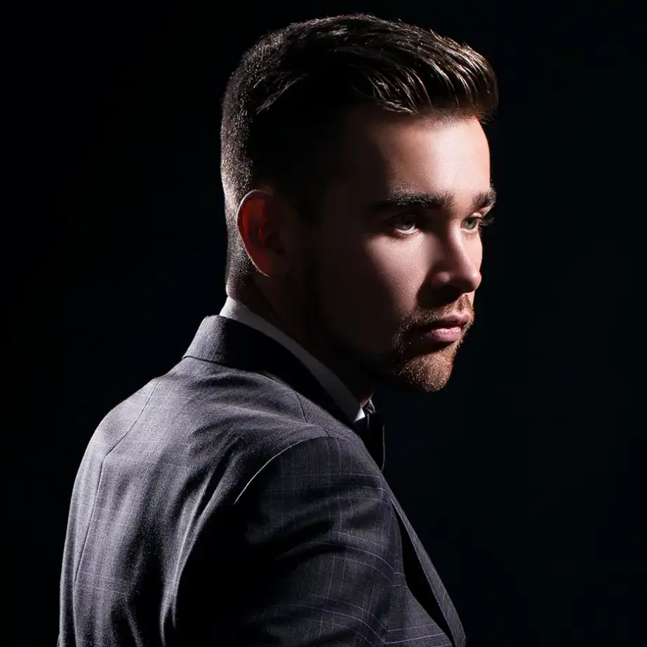 A young man in a stylish suit poses against a dark background, showcasing a confident profile. His hair is neatly styled, and he has a well-groomed beard. The dramatic lighting highlights his facial features, emphasizing a serious expression. This image conveys sophistication and professionalism, making it suitable for contexts related to fashion, business, or personal branding.