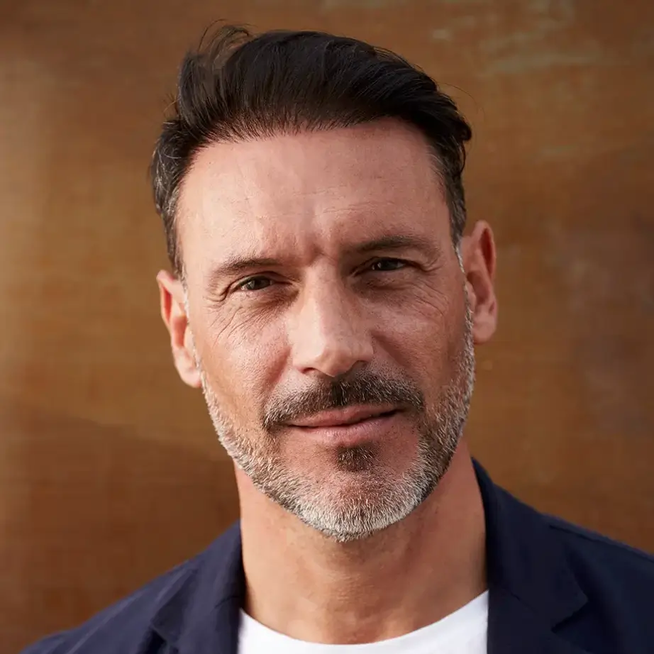 Portrait of a confident middle-aged man with short dark hair and a well-groomed beard, smiling subtly. He is wearing a navy blazer over a white t-shirt, standing against a warm-toned background. The image conveys a professional yet approachable demeanor, suitable for business or personal branding contexts. This visual representation can enhance user engagement on websites or social media platforms, particularly in industries related to lifestyle, fashion, or personal development.