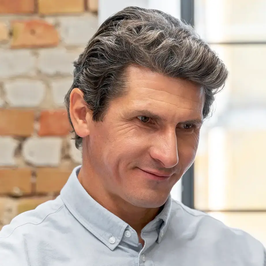 A middle-aged man with short, slightly wavy gray hair is seated in a modern office environment. He is wearing a light blue button-up shirt and appears to be engaged in a thoughtful conversation or discussion. The background features a textured wall, adding to the contemporary aesthetic of the workspace. This image conveys professionalism and focus, suitable for business-related content or articles about teamwork and collaboration.