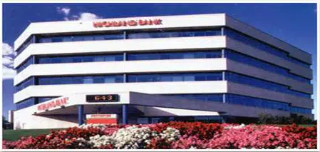 Modern multi-story building of a financial institution, featuring a prominent sign on the facade. The structure has large windows and a clean, white exterior, surrounded by vibrant flower beds in the foreground. The setting conveys a professional and welcoming atmosphere, ideal for banking services. The image highlights the architectural design and landscaping, emphasizing the institution's commitment to customer service and community engagement.
