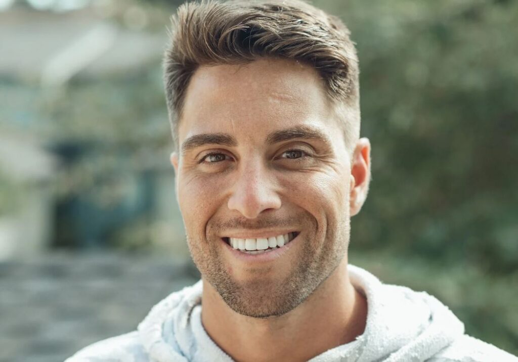 Smiling man with short, styled hair and a well-groomed beard, wearing a light hoodie, stands in front of a blurred outdoor background. His bright smile and confident expression convey a sense of warmth and approachability, making the image suitable for lifestyle or personal branding content. The natural light enhances his features, showcasing a healthy complexion. This image can be used in contexts related to positivity, health, or personal development.